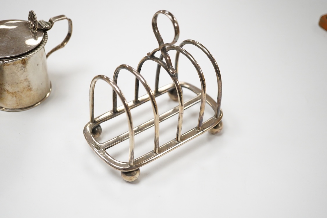An Edwardian silver mug, on claw and hoof feet, William Hutton & Sons, London 1909, height 83mm, together with a silver butter shell, mustard pot and a silver five bar toastrack, 17oz.